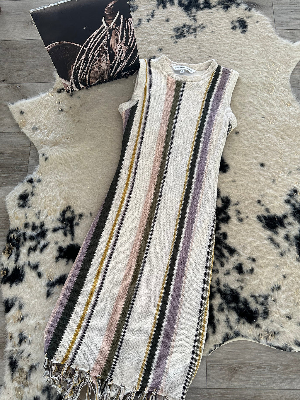A sleeveless knit dress with vertical stripes in cream, olive green, dusty pink, and mauve, featuring a fringed hem, laid flat on a black and white cowhide rug over a wood floor.