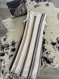 A sleeveless knit dress with vertical stripes in cream, olive green, dusty pink, and mauve, featuring a fringed hem, laid flat on a black and white cowhide rug over a wood floor.